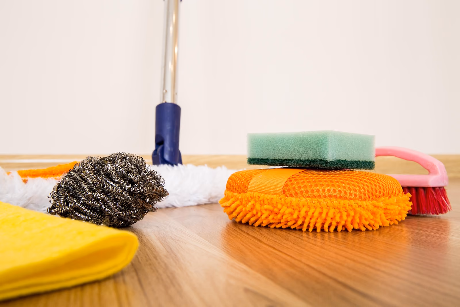 Cleaning supplies on a wooden floor: a mop, a scrubbing sponge, a microfibre mitt, a scrub brush and a yellow cloth